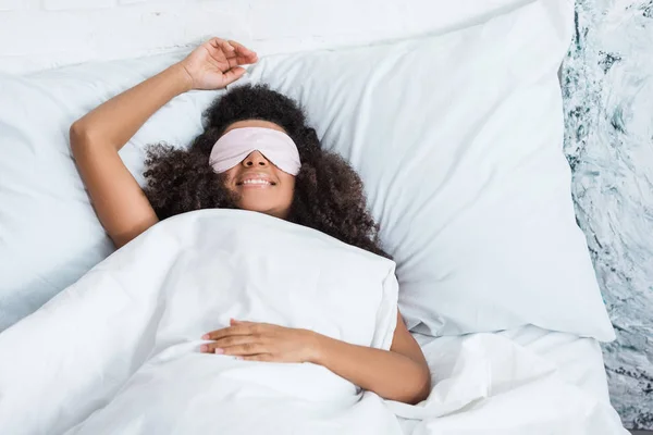 Smiling african american girl with eyes covered by sleeping blindfold in bed during morning time at home — Stock Photo