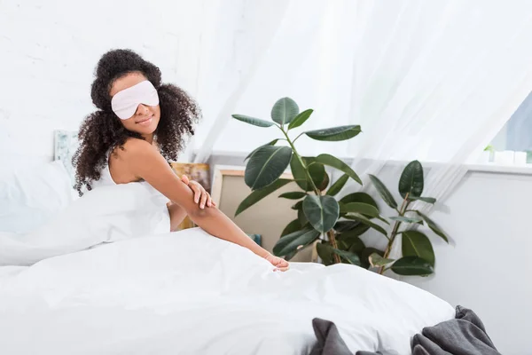 Alegre joven afroamericana chica con los ojos cubiertos por dormir venda en la cama durante la mañana en casa - foto de stock