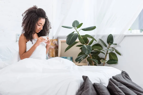 Linda menina americana africana encaracolado segurando xícara de café na cama durante o tempo da manhã em casa — Fotografia de Stock