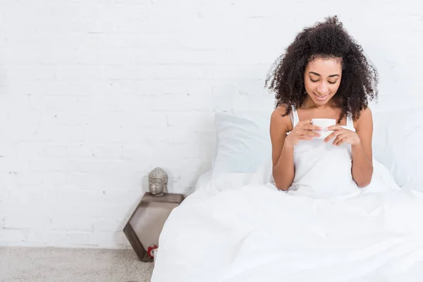 Happy curly african american girl holding cup of coffee in bed during morning time at home — Stock Photo