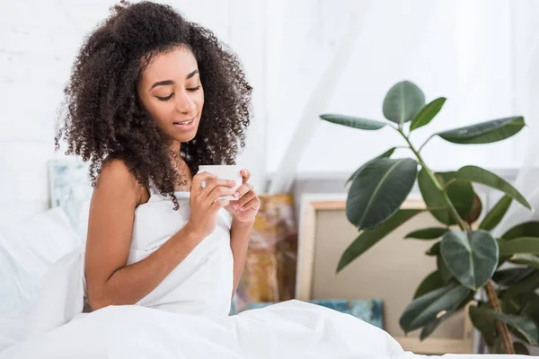 Hermosa chica afroamericana rizada sosteniendo taza de café en la cama durante la mañana en casa - foto de stock