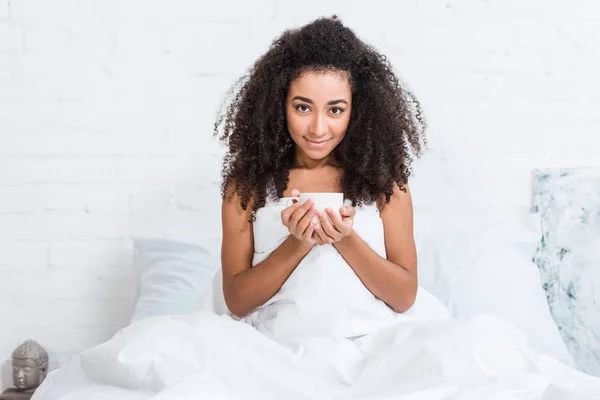 Jolie frisée afro-américaine fille tenant tasse de café au lit pendant le matin à la maison — Photo de stock