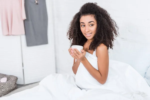 Hermosa chica afroamericana rizada sosteniendo taza de café en la cama durante la mañana en casa - foto de stock