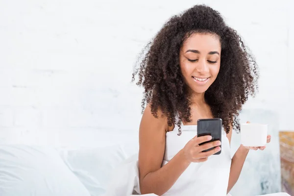 Atraente afro-americano jovem mulher com xícara de café usando smartphone na cama durante o horário da manhã em casa — Fotografia de Stock