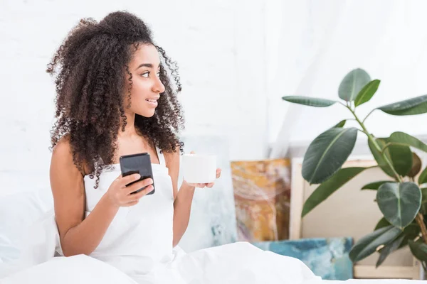 Mujer joven afroamericana con taza de café usando teléfono inteligente en la cama durante la mañana en casa - foto de stock