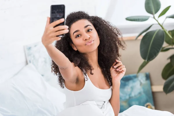 Африканская американская девушка делает утиное лицо и делает селфи на смартфоне в постели в утреннее время дома — стоковое фото