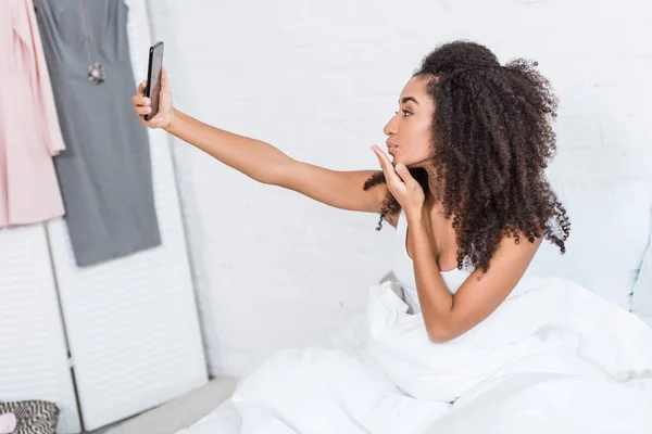 Seitenansicht eines afrikanisch-amerikanischen Mädchens, das Luftkuss pustet und morgens zu Hause ein Selfie mit dem Smartphone im Bett macht — Stockfoto