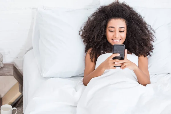 Радісна афроамериканська молода жінка використовує смартфон в ліжку в ранковий час вдома — стокове фото