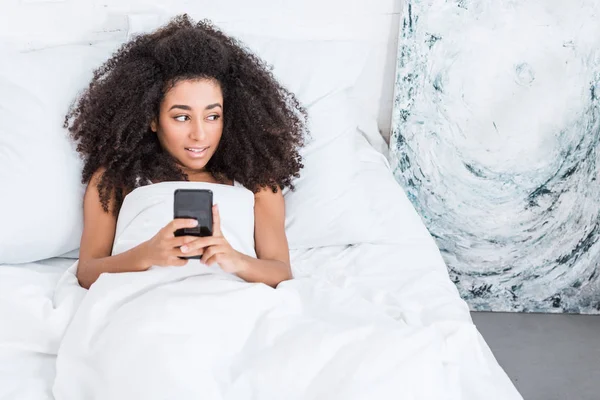 Blick aus der Vogelperspektive auf eine junge Afrikanerin, die ihr Smartphone morgens zu Hause im Bett benutzt — Stockfoto