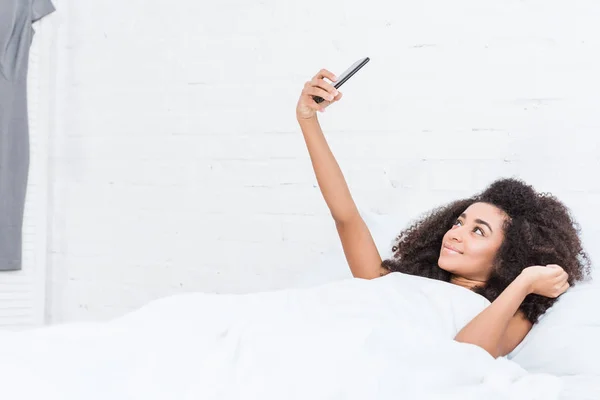 Vista lateral da menina americana africana feliz tomando selfie no smartphone na cama durante o tempo da manhã em casa — Fotografia de Stock