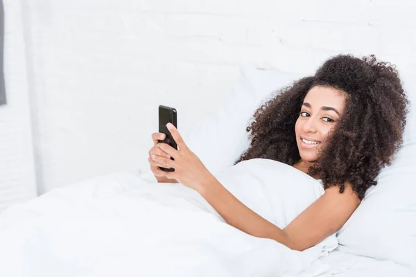 Positive afrikanisch-amerikanische junge Frau, die ihr Smartphone morgens zu Hause im Bett benutzt — Stockfoto