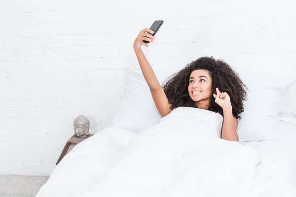 Schöne afrikanisch-amerikanische Mädchen zeigt Friedenszeichen und macht Selfie auf dem Smartphone im Bett während der Morgenzeit zu Hause — Stockfoto