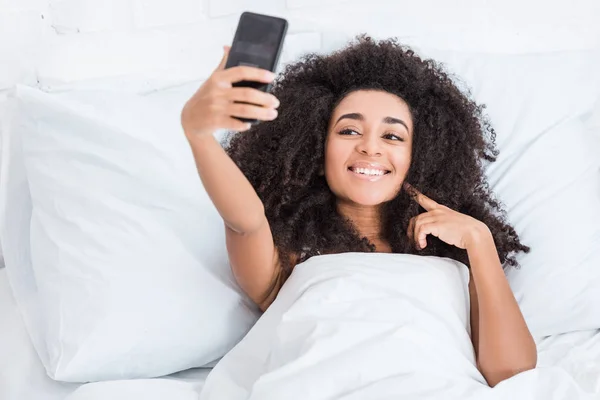 Frisé heureux afro-américain fille prendre selfie sur smartphone dans le lit pendant le matin à la maison — Photo de stock
