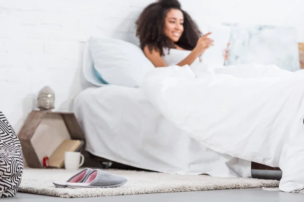 Foyer sélectif de pantoufles sur le sol et femme afro-américaine en utilisant une tablette numérique dans le lit derrière à la maison — Photo de stock