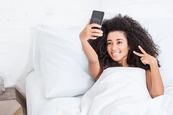 Mujer afroamericana rizada haciendo signo de victoria y tomando selfie en teléfono inteligente - foto de stock