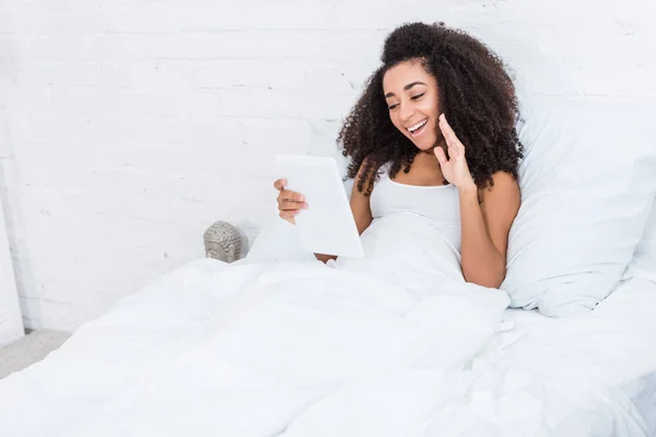 Mujer afroamericana feliz teniendo videollamada en tableta digital y saludando a mano en la cama en casa - foto de stock