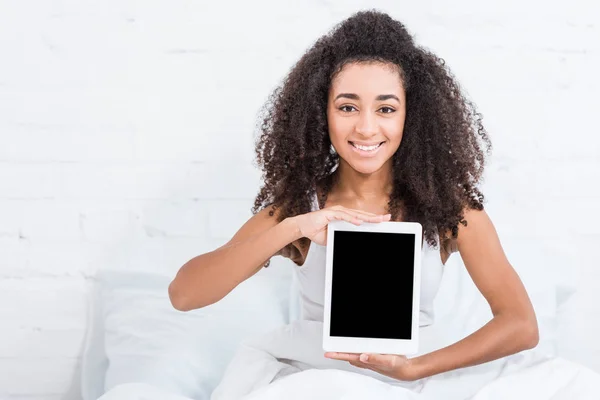 Femme afro-américaine souriante montrant tablette numérique avec écran blanc dans le lit à la maison — Photo de stock