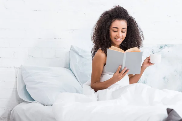 Fröhliches afrikanisch-amerikanisches Mädchen mit Kaffeetasse und Buch im Bett während der Morgenstunde zu Hause — Stockfoto