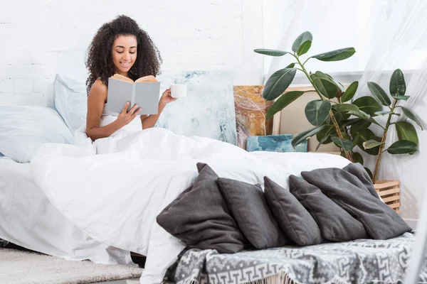 Encaracolado menina americana africana segurando xícara de café e livro de leitura na cama durante o tempo da manhã em casa — Fotografia de Stock