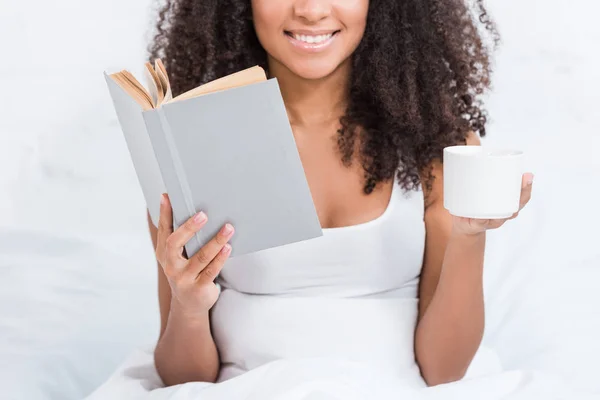 Vue partielle de fille afro-américaine tenant tasse de café et livre de lecture au lit pendant le matin à la maison — Photo de stock