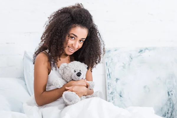 Bella sorridente ragazza riccia africana americana sdraiata a letto con orsacchiotto a casa — Foto stock
