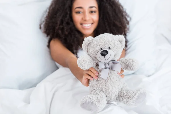 Selective focus of african american girl laying in bed and showing teddy bear at home — Stock Photo