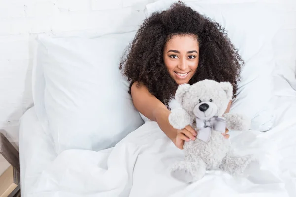 Vista ad alto angolo di giovane donna afroamericana sdraiata a letto e mostrando orsacchiotto a casa — Foto stock