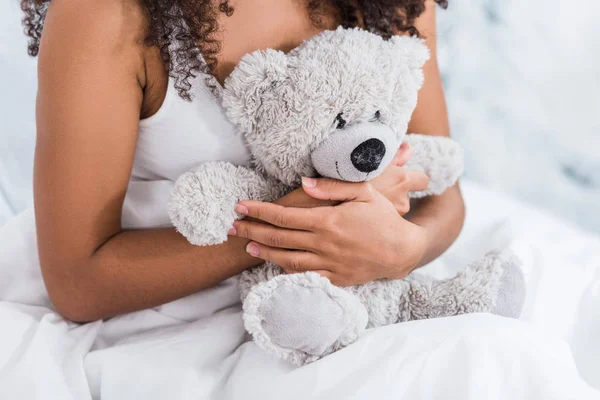 Imagen recortada de mujer joven sosteniendo oso de peluche en la cama en casa - foto de stock