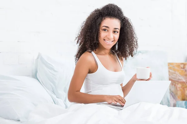 Menina encaracolado alegre segurando xícara de café e usando laptop na cama durante o tempo da manhã em casa — Fotografia de Stock