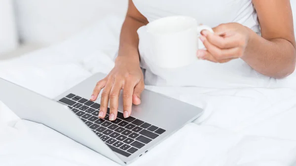 Imagen recortada de la mujer con taza de café usando el ordenador portátil en la cama durante el tiempo de la mañana en casa - foto de stock