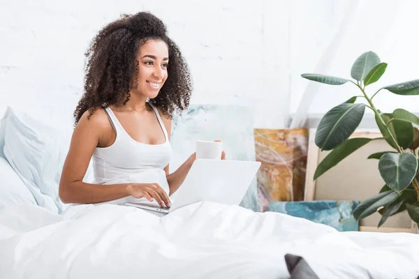 Young woman holding coffee cup and using laptop in bed during morning time at home — Stock Photo