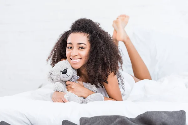 Fröhliche afrikanisch-amerikanische Mädchen liegt im Bett mit Teddybär zu Hause — Stockfoto