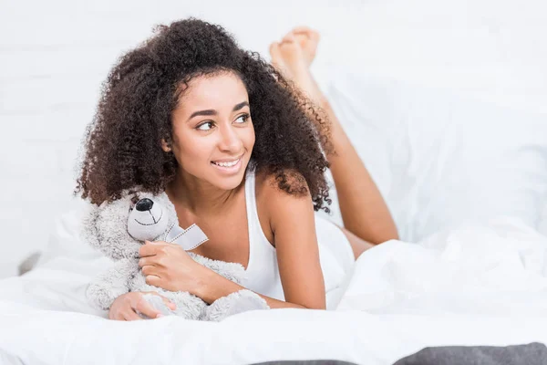 Sorridente afro-americano encaracolado menina deitado na cama com ursinho de pelúcia em casa — Fotografia de Stock
