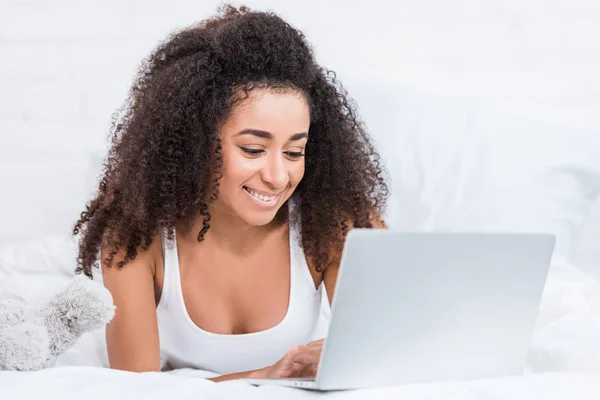 Selective focus of african american curly girl using laptop in bed during morning time at home — Stock Photo