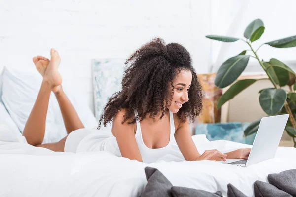 Cheerful curly girl using laptop in bed during morning time at home — Stock Photo