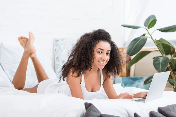 Happy african american curly girl using laptop in bed during morning time at home — Stock Photo