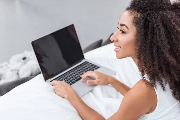Vista lateral de chica rizada afroamericana usando computadora portátil con pantalla en blanco en la cama durante el horario de la mañana en casa - foto de stock