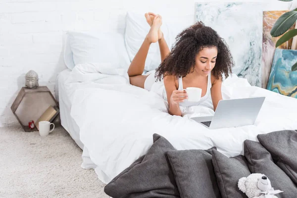 Africana chica rizada americana sosteniendo la taza de café y el uso de la computadora portátil en la cama durante la mañana en casa - foto de stock