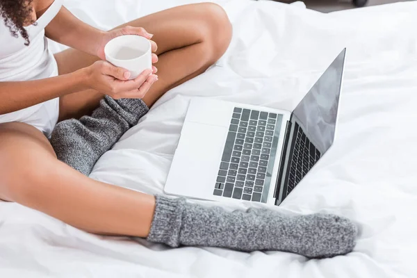 Imagen recortada de la mujer sosteniendo la taza de café y el uso de la computadora portátil con pantalla en blanco en la cama durante la mañana en casa - foto de stock