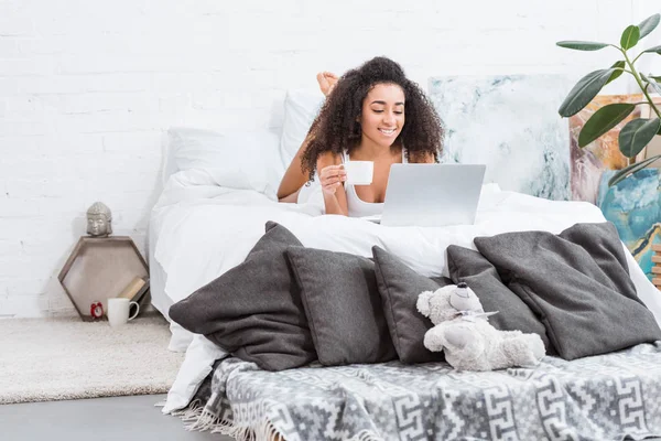 Attractive happy girl holding coffee cup and using laptop in bed during morning time at home — Stock Photo