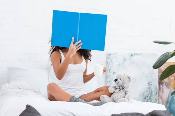 Жінка з затьмареним обличчям читає книгу і тримає чашку кави в ліжку з плюшевим ведмедем вдома — стокове фото