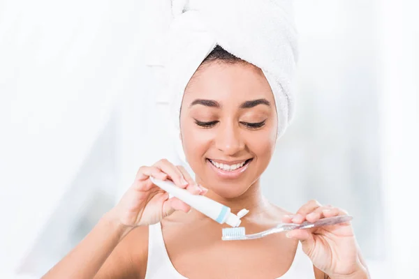 Mujer joven afroamericana con toalla envuelta en la cabeza poniendo pasta de dientes en el cepillo de dientes - foto de stock