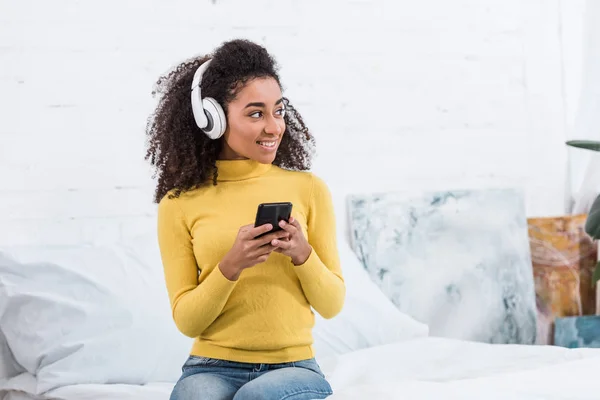 Joven afroamericana mujer en auriculares escuchando música con teléfono inteligente en casa - foto de stock