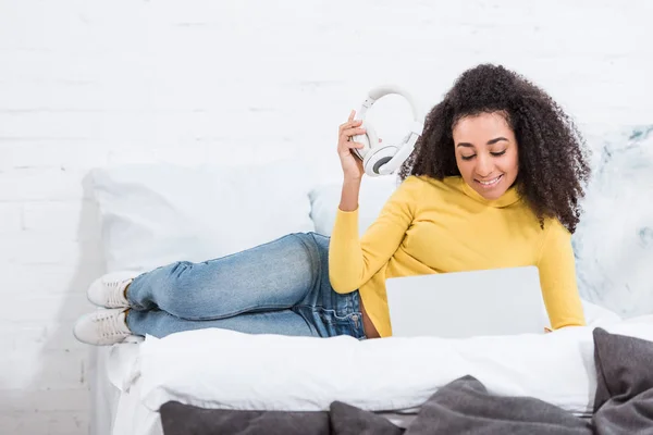 Hermosa afroamericana freelancer mujer sosteniendo auriculares y trabajando en el ordenador portátil en la cama en casa - foto de stock