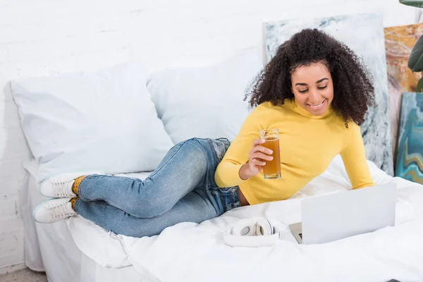 Freelancer femenina con vaso de jugo fresco trabajando en el portátil en la cama en casa - foto de stock