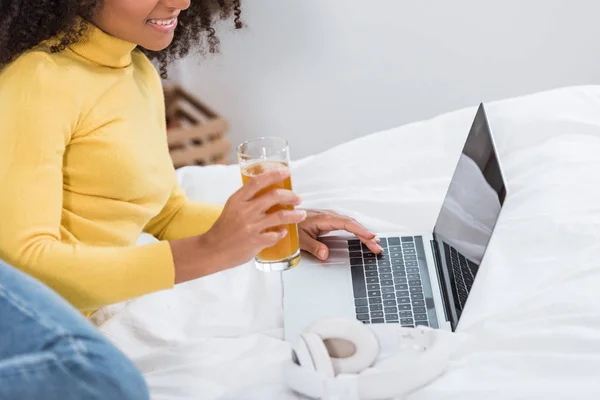 Vista parcial del freelancer femenino con vaso de jugo fresco trabajando en el ordenador portátil en la cama en casa - foto de stock