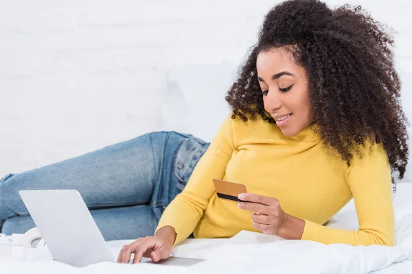 Jeune femme afro-américaine attrayante tenant la carte de crédit et faisant des achats en ligne sur ordinateur portable dans le lit à la maison — Photo de stock
