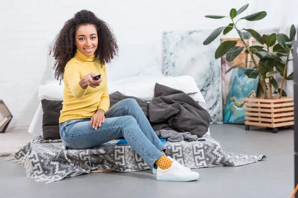 Alegre elegante afroamericana chica usando mando a distancia y viendo la televisión en casa - foto de stock