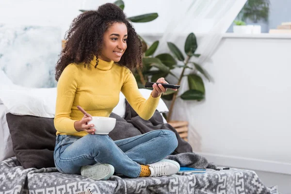 Привлекательная стильная африканская американская девушка держит миску с едой и смотрит телевизор дома — стоковое фото