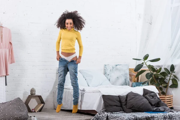Pretty african american girl putting on jeans in bedroom at home — Stock Photo